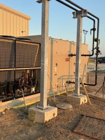 Large outdoor commercial HVAC unit with visible pipes, metal supports, and construction materials around the installation site.