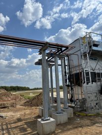 Elevated metal pipe supports and conduit system next to a large industrial cooling unit on an outdoor construction site.