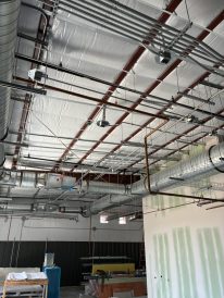 Interior view of an unfinished building with HVAC ducts, electrical conduits, insulation on the ceiling, and construction materials on the floor.